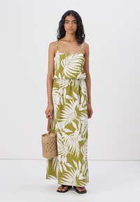 Robe maxi sans manches en vert olive avec un motif de feuilles blanches. Dispose d'une taille élastique et de bretelles réglables. Portée avec un sac en osier.