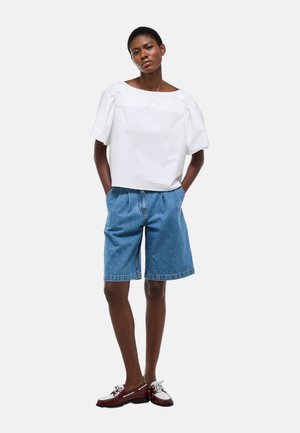 Jeune personne debout les mains dans les poches, portant un haut blanc à manches courtes, un short en jean bleu et des chaussures rouges et blanches.