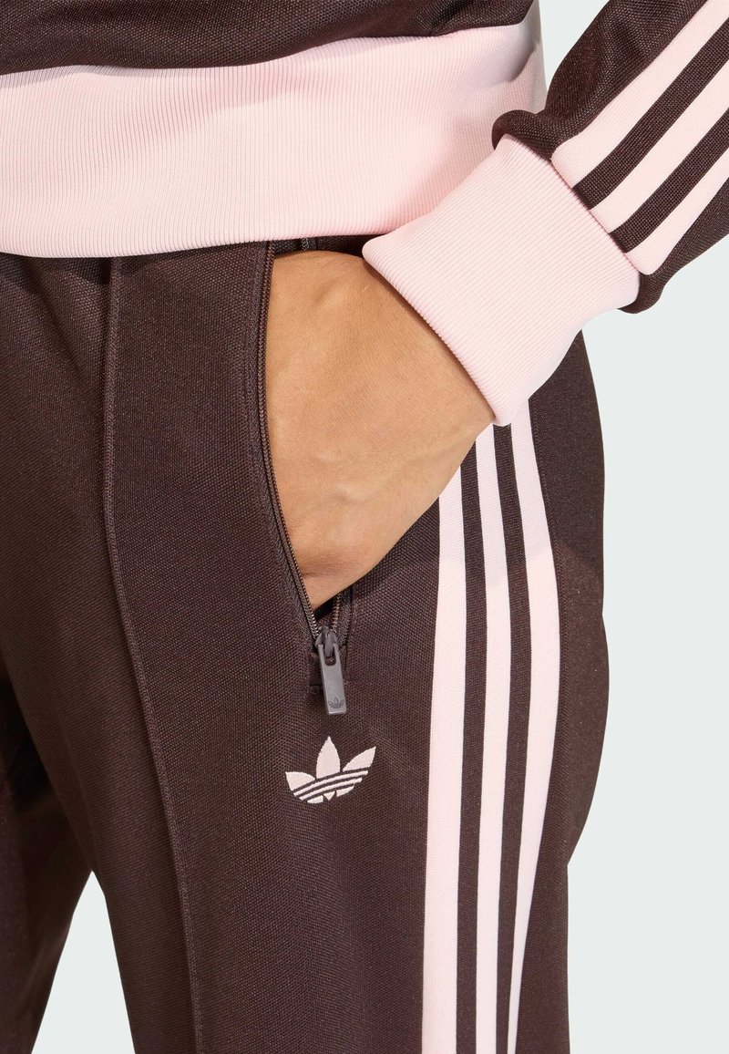 Brown athletic tracksuit pants with pink ribbed cuffs, featuring a zippered pocket and triple white stripes down the side; logo on pocket area.