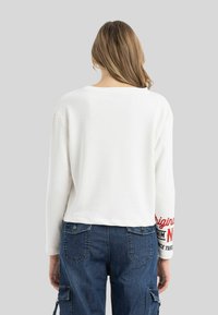 Weißer, kurzer Sweatshirt mit langen Ärmeln, mit rotem Text auf dem rechten Ärmel. Kombiniert mit blauen Denim-Cargo-Hosen, die Taschen zeigen.