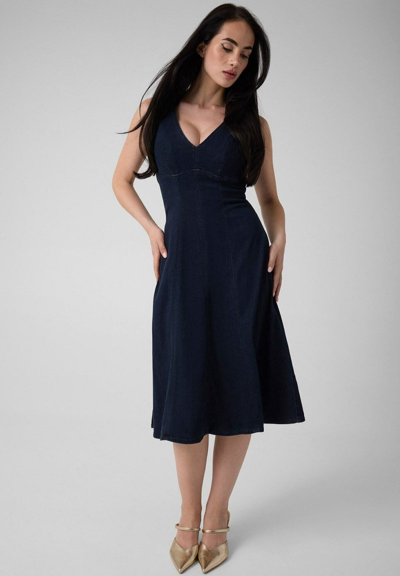 Femme aux longs cheveux foncés portant une robe midi bleu marine sans manches et des talons dorés à bout pointu, debout devant un fond clair uni.