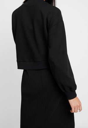 Woman wearing a black cropped sweater with ribbed cuffs and a black ribbed skirt, shown from the back against a plain background.
