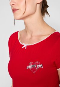 Rød bomulls T-skjorte med et hjerteformet "tommy girl"-grafikk, hvit blondedetalj og en liten hvit sløyfe ved halsen.