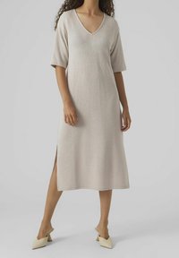 V-kaula, polvipituinen mekko vaaleanbeige värissä, jossa on lyhyet hihat ja sivurei'ät. Teksturoitu kangas; päällä vaaleat mules-kengät.