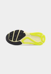 Semelle d'une basket avec des motifs de bande de roulement en caoutchouc noir et jaune néon sur une base blanche, présentée sur un fond gris clair uni.