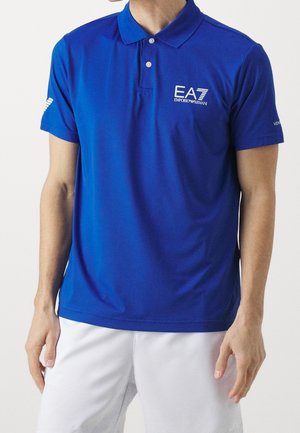 Homme portant un polo bleu Emporio Armani EA7 avec des logos blancs sur la poitrine et la manche, associé à un short blanc.
