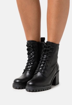 Bottes à lacets en cuir noir, montant à la cheville, avec une semelle à talon épais, une surface texturée et des œillets métalliques. Équipées d'une languette pour les tirer à l'arrière.