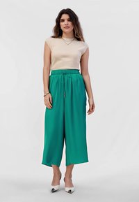 Vrouw staat met een mouwloos beige top, hoge taille wijde turquoise broek, witte spitse hakken en zilveren sieraden tegen een witte achtergrond.