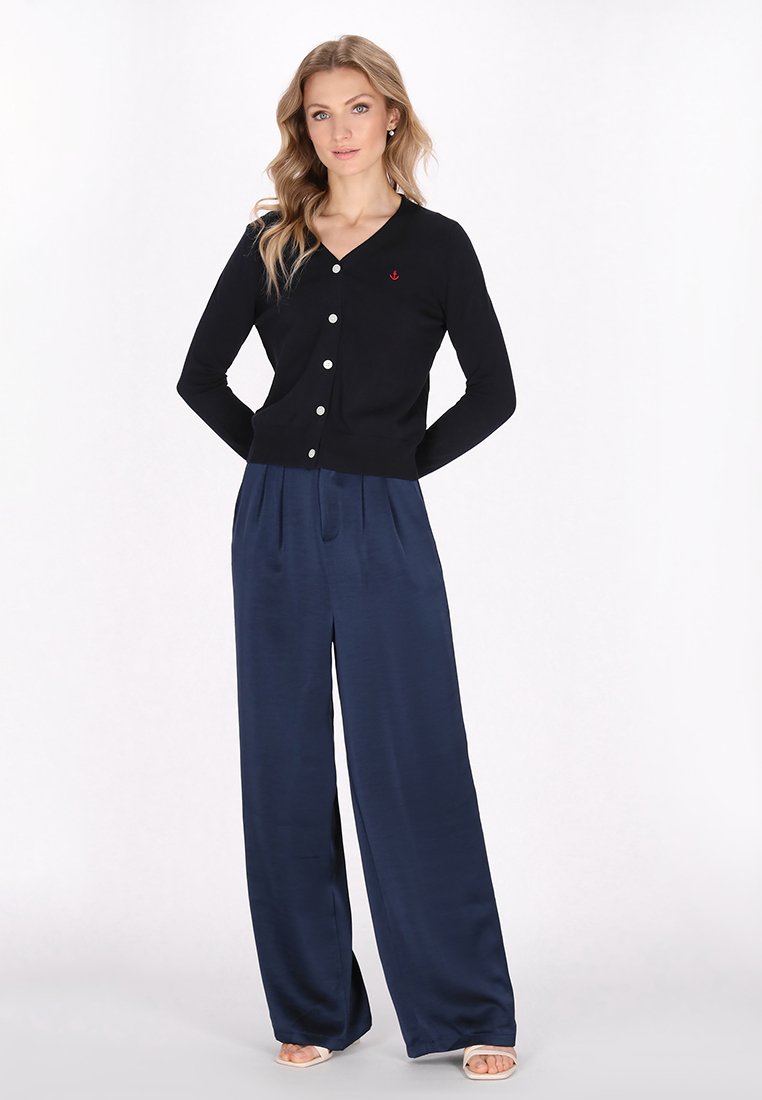 Donna con lunghi capelli ondulati, indossa un cardigan nero con bottoni e un emblema di ancora rossa, e pantaloni larghi in satin blu navy, in piedi con le mani dietro la schiena.