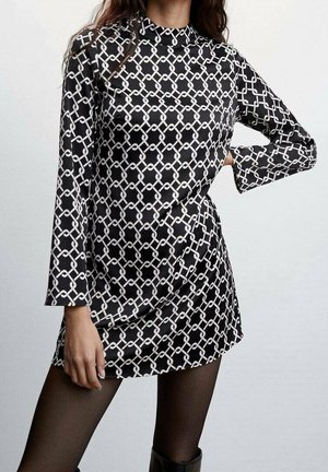 Kvinde iført en sort minikjole med hvide geometriske kædemønstre, lange ærmer og sorte strømpebukser, poserer med den ene hånd på hoften.