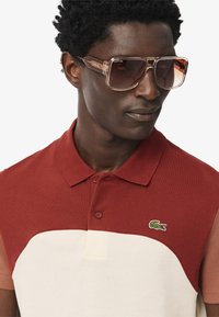 Röd och krämfärgad polo tröja med färgblockering i en texturerad tygkvalitet, som har en krage med två knappar och en liten broderad logotyp på bröstet.