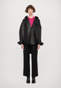 Jachetă oversized din piele neagră, cu două buzunare frontale, purtată împreună cu un tricou polo roz, pantaloni negri și botine negre.