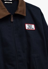 Granatowa kurtka zapinana na zamek z brązowym kołnierzem kordowym, z haftowaną naszywką, na której widnieje napis "CIESZ SIĘ WAKACJAMI NA FALACH" w białym i czerwonym kolorze.