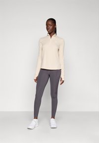 Haut zippé beige à manches longues avec un col montant, associé à des leggings gris foncé. Des baskets blanches complètent la tenue sportive. Texture lisse ; design minimaliste.