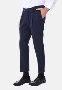 Pantaloni su misura blu navy con pince frontali, tasche a taglio e chiusura con bottone. Indossati con mocassini neri e calzini neri.