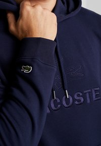 Marineblå hættetrøje lavet af blødt stof. Har en broderet logo og en lille grøn krokodille-patch på ærmet. Inkluderer snoretræk-detaljer.