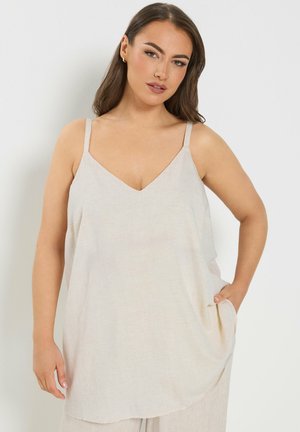 Vrouw met lang bruin haar, gekleed in een mouwloze beige top en bijpassende broek, staand met één hand in de zak tegen een witte achtergrond.