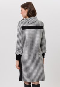 Grijs en zwarte jurk met een geometrisch patroon, featuring een brede kraag, lange mouwen, zijpanelen en een accentstreep op de rug.