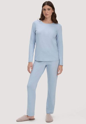 Frau trägt hellblaues Langarmoberteil und Hose, steht entspannt mit beigen Hausschuhen vor einem schlichten hellen Hintergrund.