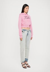 Hoodie corto rosa con testo nero, abbinato a jeans azzurri a vestibilità rilassata. Il modello indossa sandali neri con tacco e cinturini alla caviglia.