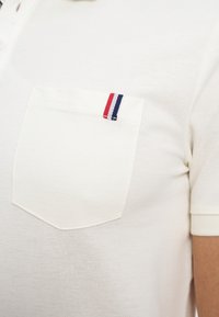 Chemise blanche à manches courtes avec une poche sur la poitrine gauche, comportant une petite étiquette rayée rouge, blanche et bleue.