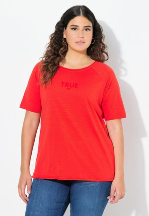 T-shirt rouge à manches courtes avec un col rond et l'inscription "TAILLE RÉELLE" imprimée en texte foncé au centre. Texture lisse ; coupe décontractée.