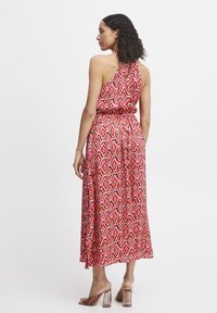 Maxi dress rosso con motivo, collo halter, vita elastica e orlo diagonale. Il tessuto presenta forme geometriche e una texture liscia.