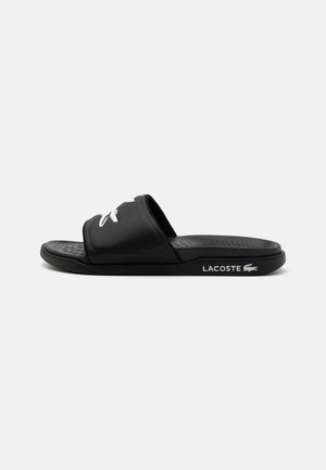 Black slide sandal made of rubber, featuring a textured footbed, two wide straps, and a white Lacoste logo on the side.