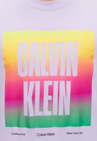 Λευκό πουκάμισο με μεγάλο κείμενο CALVIN KLEIN πάνω σε κίτρινο, ροζ και πράσινο τετράγωνο με διαβάθμιση και μικρότερο κείμενο που αναγράφει Established 1968, Calvin Klein, New York City.