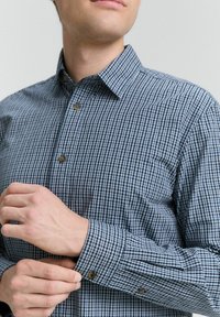 Camisa de cuadro azul de botones con cuello abierto, que presenta pequeños botones marrones y un bolsillo en el pecho, fabricada con una tela ligera.