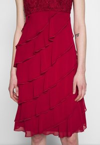 Czerwona sukienka z dopasowaną koronkową górą i warstwową spódnicą z falbanami. Materiał wydaje się lekki i zwiewny, charakteryzuje się gładką fakturą i subtelnym połyskiem.