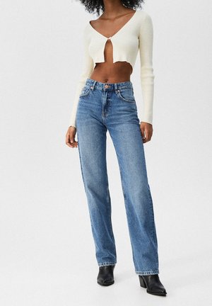 Kermanvärinen, ribbikuvioitu, pitkähihainen lyhyt neule V-kaula-aukolla, yhdistetty vaaleansinisiin korkeavyötäröisiin suoralahkeisiin farkkuihin ja mustiin korkokengillisiin saappaisiin.