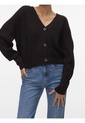 Personne portant un cardigan en maille noir boutonné et un jean bleu, posant avec une main sur la hanche et l'autre reposant sur la cuisse.