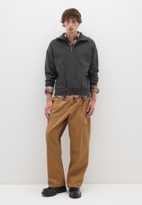 Grauer Pullover mit halbem Reißverschluss; darunter ein braunes kariertes Hemd; locker sitzende sandfarbene Cargo-Shorts mit Seitentaschen; schwarze klobige Schuhe.