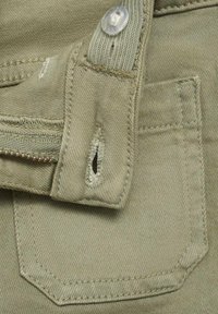 Tissu khaki avec une fermeture éclair et une petite poche de forme arrondie, coutures visibles et un détail de trou usé.