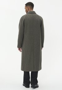 Cappotto lungo a motivo houndstooth in verde oliva con colletto strutturato e maniche raglan; indossato sopra pantaloni scuri e scarpe.