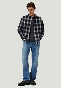 Ung man med händerna i fickorna, iklädd en svartvit rutiga skjorta, mörk t-shirt, blå jeans med slitna detaljer och svarta skor.