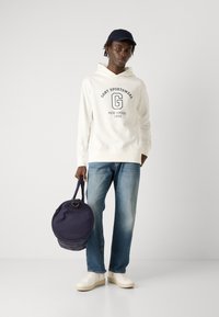 Sudadera blanca con capucha y estampado 'GANT SPORTSWEAR', jeans de mezclilla azul, zapatillas blancas y un bolso redondo azul marino con asa.