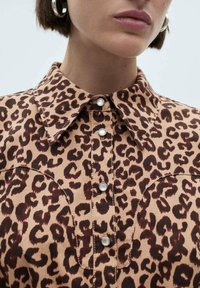 Halenka s leopardím vzorem na béžovém podkladu, s hnědými tečkami, špičatým límcem a detaily s knoflíky. Materiál se zdá být tkanina.