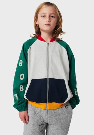 Kind met schouderlang blond haar, gekleed in een groene, grijze, marineblauwe en gele ritsjack met tekst op de mouwen en grijze gewatteerde broek.