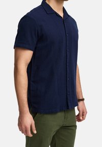 Uomo che indossa una camicia a maniche corte blu navy con bottoni e pantaloni verde oliva, in piedi con le braccia rilassate ai lati.