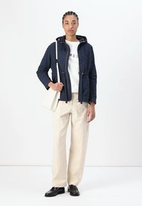 Ναυτικό γεμιστό μπουφάν με ρυθμιζόμενη θηλιά, λευκή μπλούζα με λογότυπο, κρεμ παντελόνι και μαύρες loafers. Μινιμαλιστικός σχεδιασμός με διακριτά μοτίβα.