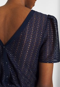 Haut bleu marine à motif en tissu transparent texturé, avec un col en V et des manches courtes bouffantes. Fermeture par bouton au dos.