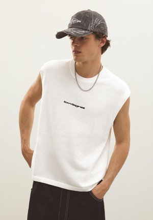 Jeune homme portant un haut en maille blanc sans manches, une casquette noire avec des motifs blancs, un collier chaîne argenté et un pantalon noir avec des coutures blanches.