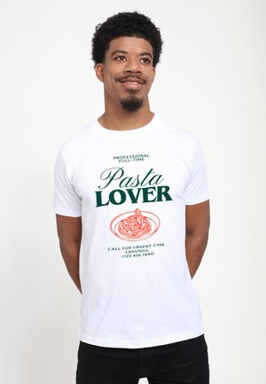 Homme souriant portant un T-shirt blanc avec le texte "Professional Full-Time Pasta Lover" et un graphique de bol de pâtes rouge, debout devant un fond uni.