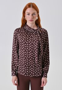 Una donna con lunghi capelli rossi indossa una camicia marrone a pois con una cravatta e maniche lunghe, abbinata a pantaloni marroni su uno sfondo bianco.