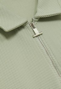 Cămașă polo verde deschis, dintr-un material texturat, cu închidere prin fermoar și guler pliat. Un model geometric subtil adaugă detalii.