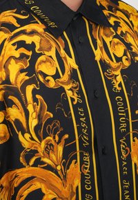 Camicia a maniche corte nera con intricate fantasie floreali dorate e bold strisce gialle, con il testo "VERSACE COUTURE". Stile classico con colletto.