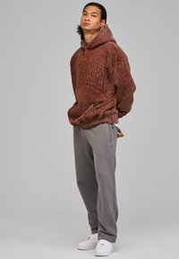 Hoodie marrone sbiadito con lettering in rilievo, abbinato a pantaloni della tuta grigi e sneaker bianche. Tessuto morbido e vestibilità rilassata.