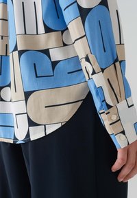 Camicia con colletto caratterizzata da una stampa geometrica in blu, beige e nero. Realizzata in tessuto morbido. Presenta polsini con bottone e un orlo curvo.